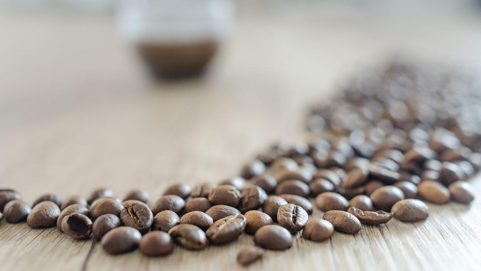 Whole roasted coffee beans scattered across a light wooden surface in bright daylight