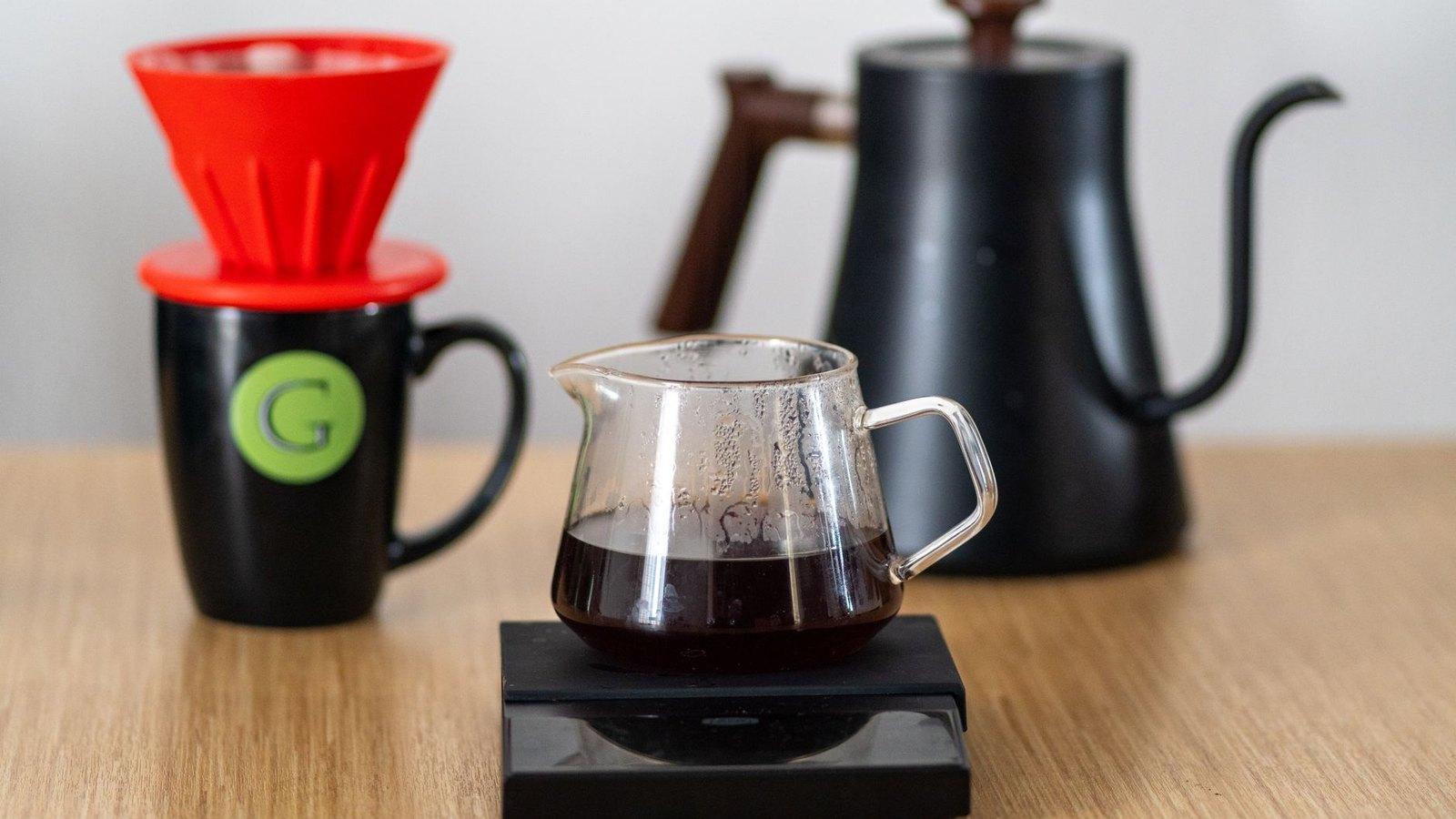 A pour over setup on a wooden table: a red V60 dripper, a glass carafe on a scale, and a gooseneck kettle