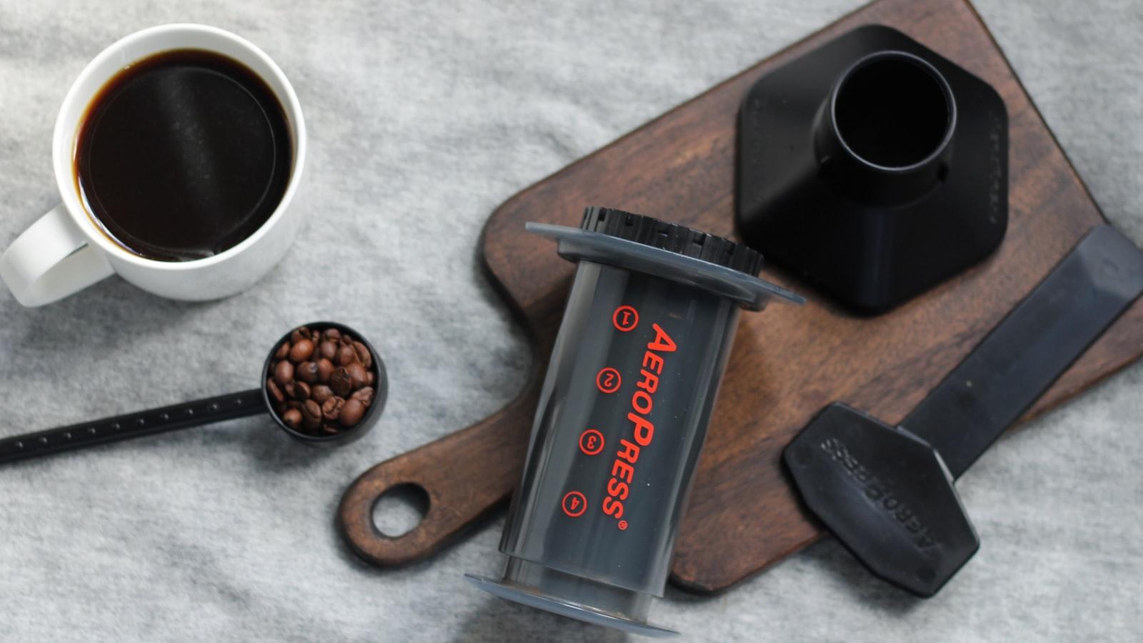 A disassembled AeroPress laid out on a wooden board: the clear chamber, the filter cap, a scoop of beans, the stirrer paddle and a mug of black coffee
